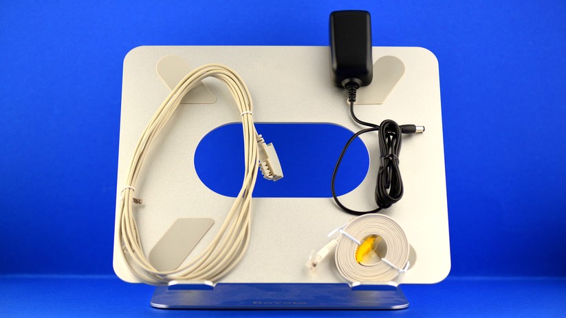 Der restliche Lieferumfang hält sich in Grenzen. Links außen ein hellbeiges DSL-Kabel. Rechts unten ein weißes Ethernet-LAN-Kabel, nicht rund, sondern flach. Das lässt sich relativ unauffällig verlegen: Hinter Fußbodenleisten, auf weißer Wand, auf weißem Fensterrahmen.