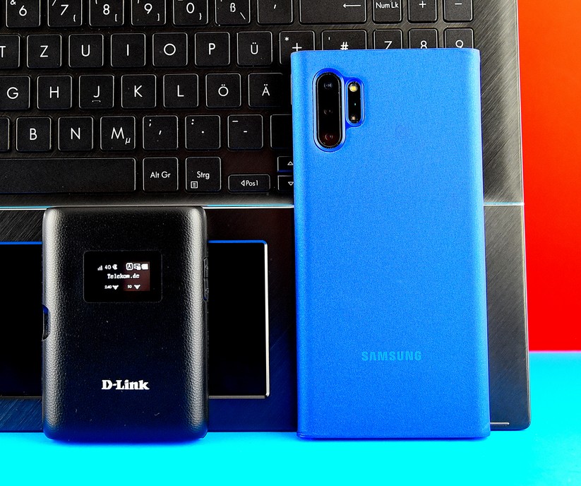 Der schwarze D-Link DWR-933 Router links unten holt das Internet per LTE bis 300 MBit/s aus der Mobilfunkluft und funkt es per WiFi-5 an unseren WLAN-schnellsten (aber LTE-losen) Laptop Asus ZenBook Flip 15 mit WiFi-6 bis 2400 MBit/s weiter. Das blaue Samsung Galaxy Note 10+ (rechts) hat ein eigenes LTE-Modul, kann aber auch über den WLAN-Hotspot des D-Link-Routers im Internet surfen (Bild: Harald Karcher).